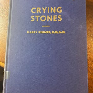 May include: A hardcover book titled "Crying Stones" with gold lettering on a blue cover. The author's name, Harry Rimmer, D.D., Sc.D., is printed below the title. The book has a textured appearance.