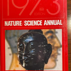 Op de afbeelding: Een rood boek met de titel "1973 TIME-LIFE BOOKS EDITION NATURE/SCIENCE ANNUAL" en een zwart-witfoto van een acupuncturist-oefenpop op de omslag. De pop is een zwarte buste met naalden die in het gezicht en hoofd zijn gestoken.