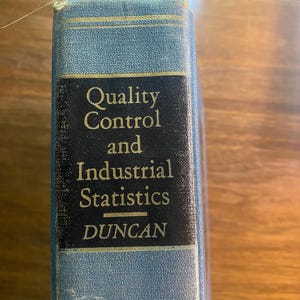 May include: A blue hardcover book spine with a black rectangular label featuring the gold-colored text Quality Control and Industrial Statistics and the author name DUNCAN. The book spine has thin gold horizontal lines above and below the label.