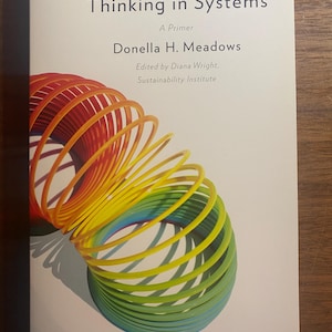 May include: A book titled Thinking in Systems: A Primer by Donella H. Meadows, edited by Diana Wright, Sustainability Institute. The cover features a colourful, coiled spring toy in shades of red, orange, yellow, green, and blue on a white background.