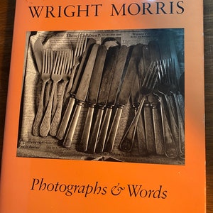May include: A book cover with an orange background and the title "WRIGHT MORRIS Photographs & Words". The cover features a black and white photograph of a collection of silverware, including forks and knives, arranged on top of a newspaper.