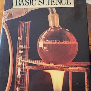 Może przedstawiać: Vintage okładka książki zatytułowana "THE MIND ALIVE ENCYCLOPEDIA BASIC SCIENCE" zawiera ilustrację eksperymentu naukowego. Okrągła kolba znajduje się nad płomieniem, z cylindrem miarowym i rurkami. Tło jest ciemne.