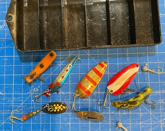 May include: A collection of fishing lures in a black plastic tackle box. The lures are various colors, including red, white, blue, yellow, and black. Some of the lures have a metallic finish. The lures are arranged in a grid pattern on a blue cutting mat.