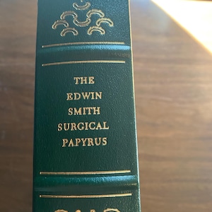 Può includere: Un libro rilegato in pelle verde con lettere dorate sul dorso. Il titolo è "The Edwin Smith Surgical Papyrus".