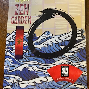 Pode incluir: Uma capa de livro com um título dourado que diz "Royal Zen Garden" e um círculo preto sobre um fundo de ondas azuis e brancas. O canto inferior direito tem um logotipo vermelho com o texto "Garden at Home" e "Your Garden Gift Specialist".