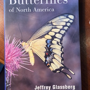 May include: Book cover featuring a yellow and black butterfly with blue and red markings, perched on a pink flower. The title "Butterflies of North America" is printed in white on a dark blue background. The author's name, "Jeffrey Glassberg", is printed in white below the butterfly. The text "Approved by the North American Butterfly Association (NABA)" is printed in white at the bottom of the cover.