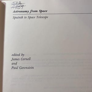 May include: A book page with the title "Astronomy from Space: Sputnik to Space Telescope" printed in black. The editors are James Cornell and Paul Gorenstein. Handwritten numbers are at the top of the page. A red stamp is visible.