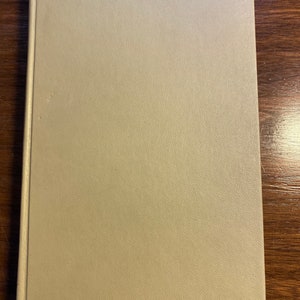 May include: A closed book with a light tan cover. The cover has a textured, slightly speckled appearance. The book is rectangular and appears to be a hardcover book. The book is lying on a dark brown wooden surface.