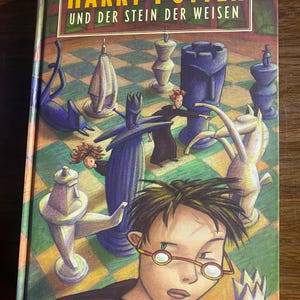 May include: A hardcover book titled Harry Potter und der Stein der Weisen by Joanne K. Rowling. The cover features a colorful illustration of a person with dark hair and glasses in the foreground, with giant chess pieces on a checkered board.