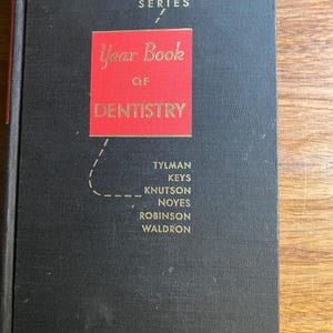 May include: A vintage black hardcover book titled "Year Book of Dentistry" with red lettering and a red square. The book is from the 1961-1962 series and lists several authors. The book is on a wooden surface.