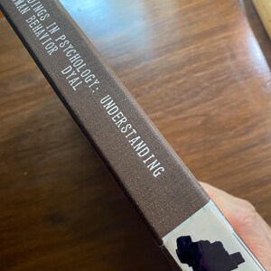 May include: A brown book spine with the title "Readings in Psychology: Understanding Human Behavior" in white text. The book has a white label with dark blue markings.