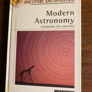 Op de afbeelding: Een boek getiteld "Modern Astronomy" met de ondertitel "Expanding the Universe". De omslag toont een silhouet van een persoon die door een telescoop kijkt tegen een achtergrond van wervelende sterren. Onderdeel van de serie "Milestones in Discovery and Invention".