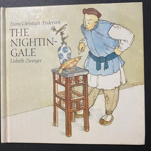Può includere: Un'illustrazione di copertina di un libro che mostra un uomo con una veste blu e bianca che indica un piccolo uccello su un tavolo. Il titolo del libro è "The Nightin-Gale" di Hans Christian Andersen.