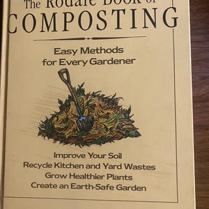 May include: A hardcover book titled The Rodale Book of Composting, New, Revised Edition. The cream-colored cover features a central illustration of a blue garden fork in a pile of brown and green compost, with text detailing methods for gardeners.