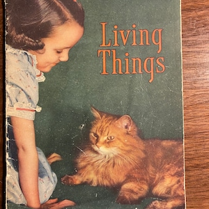 May include: A vintage book titled "Living Things" with a green cover. The cover features a young girl with a red hair clip looking down at a ginger cat.