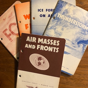 Puede incluir: Una pila de cuatro libros de bolsillo vintage con cubiertas marrones, azules y rosas. Las cubiertas presentan texto e ilustraciones relacionadas con el clima y la meteorología. El texto en la portada del libro inferior dice "Air Masses and Fronts" y "Aeralogy Series Number Four".