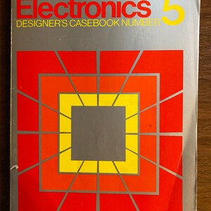 Può includere: Una copertina argentata di un libro intitolato "Electronics 5" con le parole "Designer's Casebook Number" stampate sotto il titolo. La copertina presenta un design geometrico con un quadrato centrale grigio, circondato da quadrati concentrici gialli e rossi.