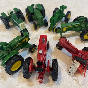 May include: A collection of toy tractors in various shades of green and red. The tractors have black tires and yellow accents. One red tractor has a tag that reads "Midwest Importers".
