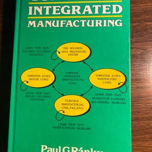 Op de afbeelding: Een groene boekomslag met de titel "Computer Integrated Manufacturing" in witte en gele letters. De boekomslag bevat ook een diagram van het computergeïntegreerde productieproces met de tekst "Learn from past business failures/successes", "The business data processing system", "Computer aided design (CAD)", "Computer integrated manufacturing (CIM)", "Computer aided manufacture (CAM)", "Flexible manufacturing (FMS, FAS, etc)", "Learn from past design failures/successes", "Learn from past production planning and control problems", en "Learn from past manufacturing problems". De naam van de auteur, "Paul GRánky", is in witte en gele letters onderaan de omslag gedrukt.