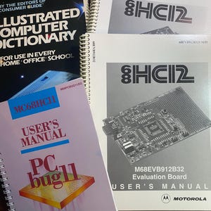 May include: A collection of computer-related manuals and a dictionary. The manuals include "MC68HC11 User's Manual" and "M68EVB912B32 Evaluation Board User's Manual" with the Motorola logo. The dictionary is titled "Illustrated Computer Dictionary".