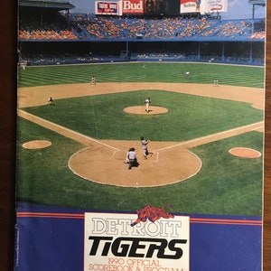 Op de afbeelding: Een blauw en oranje officieel scoreboek en programma van de Detroit Tigers uit 1990. De omslag toont een tijgerlogo en een foto van een baseballwedstrijd die gaande is.