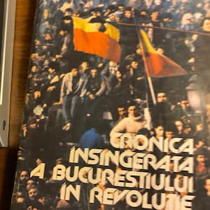May include: A vintage book cover featuring a black and white photograph of a crowd of people holding a yellow, red and blue flag. The title of the book is "Cronica Insingerata A Bucurestiului In Revolutie".
