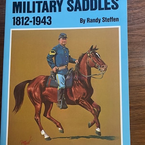 Puede incluir: Portada del libro titulada "UNITED STATES MILITARY SADDLES 1812-1943" de Randy Steffen. La portada presenta una ilustración de un soldado a caballo. El soldado lleva un uniforme azul y monta un caballo marrón.