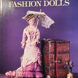 Puede incluir: Una portada de libro con el título "How to Collect French Fashion Dolls" de Mildred y Vernon Seeley. La portada presenta una muñeca de porcelana con un vestido rosa y morado, sosteniendo una sombrilla blanca y de pie junto a un baúl de madera.