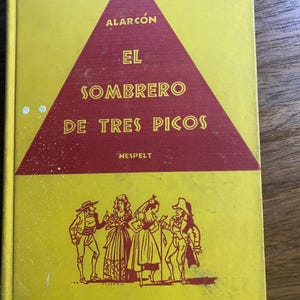 Puede incluir: Un libro antiguo con una cubierta amarilla y un triángulo rojo con el título "EL SOMBRERO DE TRES PICOS" y el nombre del autor, Alarcón. La portada también incluye ilustraciones de personas con ropa de época.