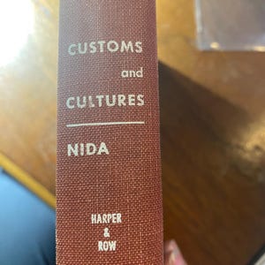 May include: A hardcover book with a textured, reddish-brown cover. The title "CUSTOMS and CULTURES" is printed in silver lettering, along with the author's name "NIDA" and the publisher "HARPER & ROW". The book's spine is visible.