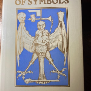 Puede incluir: Una cubierta de libro con el título "A Dictionary of Symbols" en letras doradas. La cubierta presenta una ilustración detallada de una criatura alada con cabeza humana y un ojo en el pecho, sobre un fondo azul.