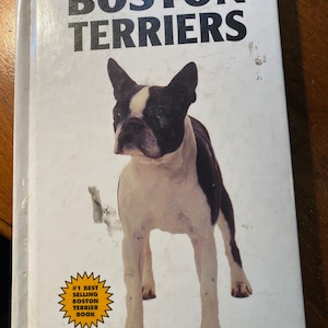 May include: A white book with the title "Boston Terriers" in black text. The cover features a black and white Boston Terrier dog standing on a white background. A yellow starburst sticker with the text "#1 Best Selling Boston Terrier Book" is in the lower left corner.