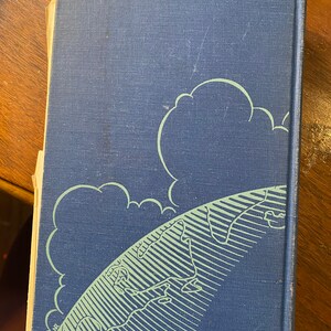 Op de afbeelding: Een blauw hardback boek met een groene en witte illustratie van een wereldbol en wolken op de omslag.