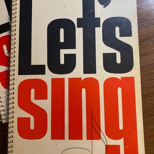 May include: A spiral-bound book with the words "Let's Sing" in large, bold, black and red letters. The cover is off-white, and the book is lying on a wooden surface. A musical note is faintly drawn on the cover.