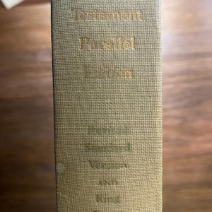 Puede incluir: Primer plano del lomo de un libro con el título "New Testament Parallel Edition" en letras doradas. El libro es de color beige e incluye la Versión Estándar y la Versión King James.