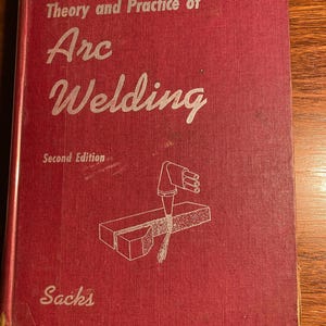 May include: A red hardcover book titled "Theory and Practice of Arc Welding" with the author's name "Sacks" and the words "Second Edition" printed on the cover. The book features a line drawing of a welding torch and a metal piece being welded.
