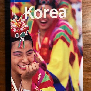 May include: A Lonely Planet travel guide for Korea with a colorful image of a woman in traditional Korean clothing smiling.