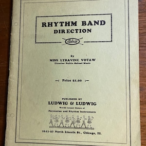 May include: A vintage book cover for "Rhythm Band Direction" by Miss Lyravine Votaw, Director of Public School Music. The cover features a black and white illustration of a marching band with percussion instruments and the Ludwig logo. The book is published by Ludwig & Ludwig, World's Largest Makers of Percussion and Rhythm Instruments, located at 1611-27 North Lincoln St., Chicago, Ill.