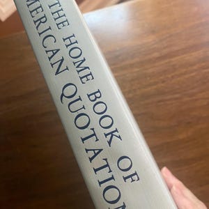 以下が含まれることがあります： 「THE HOME BOOK OF AMERICAN QUOTATIONS」というタイトルが、濃い青色の文字で、明るい灰色の背景に書かれた本の背表紙。ハードカバーのようです。