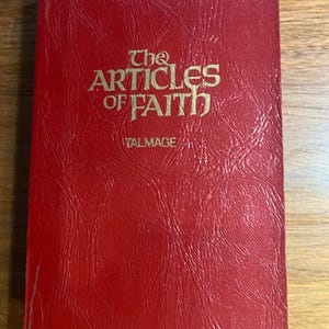 Puede incluir: Un libro rojo titulado "The Articles of Faith" con letras doradas. La portada tiene una apariencia texturizada, similar al cuero. El nombre del autor, Talmage, está impreso debajo del título.