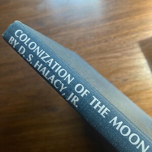Può includere: Primo piano di un libro intitolato "Colonization of the Moon" di D.S. Halacy, Jr. È visibile il dorso del libro, con testo bianco su sfondo blu strutturato. Il libro è appoggiato su una superficie di legno scuro.