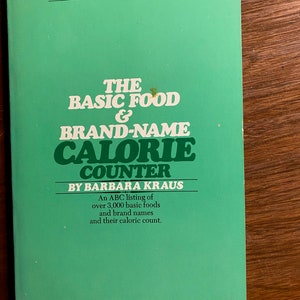 Puede incluir: Un libro de bolsillo verde titulado "THE BASIC FOOD & BRAND-NAME CALORIE COUNTER" de Barbara Kraus. El libro incluye una lista ABC de más de 3.000 alimentos básicos y nombres de marcas con su conteo calórico. La portada también presenta una cita de Women's Wear Daily.