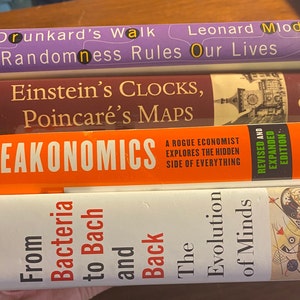 Puede incluir: Una pila de cuatro libros con cubiertas de colores. El libro de arriba es morado y tiene el título "The Drunkard's Walk: How Randomness Rules Our Lives" de Leonard Mlodinow. El segundo libro es marrón y tiene el título "Einstein's Clocks, Poincaré's Maps" de Peter Galison. El tercer libro es naranja y tiene el título "Freakonomics: A Rogue Economist Explores the Hidden Side of Everything" de Steven D. Levitt y Stephen J. Dubner. El libro de abajo es blanco y tiene el título "From Bacteria to Bach: The Evolution of Minds" de Daniel C. Dennett.