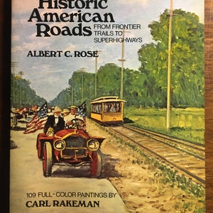 Pode incluir: Capa de livro com uma pintura de um carro antigo dirigindo em uma estrada com um bonde no fundo. O título do livro é "Historic American Roads: From Frontier Trails to Superhighways" de Albert C. Rose. A capa também indica que contém 109 pinturas a cores de Carl Rakeman.