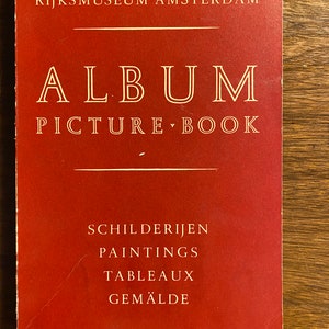 May include: A red book with the title "ALBUM PICTURE BOOK" and the text "RIJKS MUSEUM AMSTERDAM" at the top. The book also has the text "SCHILDERIJEN PAINTINGS TABLEAUX GEMÄLDE" in white.