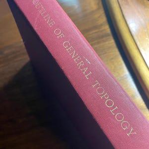 Op de afbeelding: Een bordeauxrood boek met de titel "Outline of General Topology" met gouden letters. De rug van het boek is zichtbaar en toont de titel en een gestructureerde kaft. Het boek staat schuin, wat suggereert dat het op een houten oppervlak ligt.