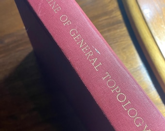 May include: A burgundy book titled "Outline of General Topology" with gold lettering. The book's spine is visible, showing the title and a textured cover. The book is angled, suggesting it's resting on a wooden surface.