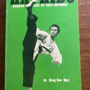 Op de afbeelding: Een vintage boek getiteld "HAPKIDO" met de ondertitel "KOREAN ART OF SELF-DEFENSE" op een groene achtergrond. De cover toont een man in een vechtsportuniform die een hoge trap uitvoert. De naam van de auteur, Bong Soo Han, is ook zichtbaar.