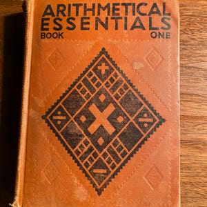 May include: A vintage brown book titled "Arithmetical Essentials Book One" with a black geometric design on the cover.