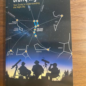 May include: A book cover with a dark blue background and a star chart showing constellations like Ursa Major, Ursa Minor, and Cassiopeia. The title of the book is "Starry Night Companion: Your Guide to Understanding the Night Sky". Three silhouettes of people are shown looking through telescopes at the night sky.